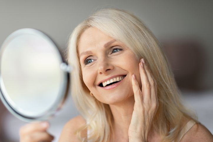 Smiling woman looking in a handheld mirror, touching her cheek in soft light