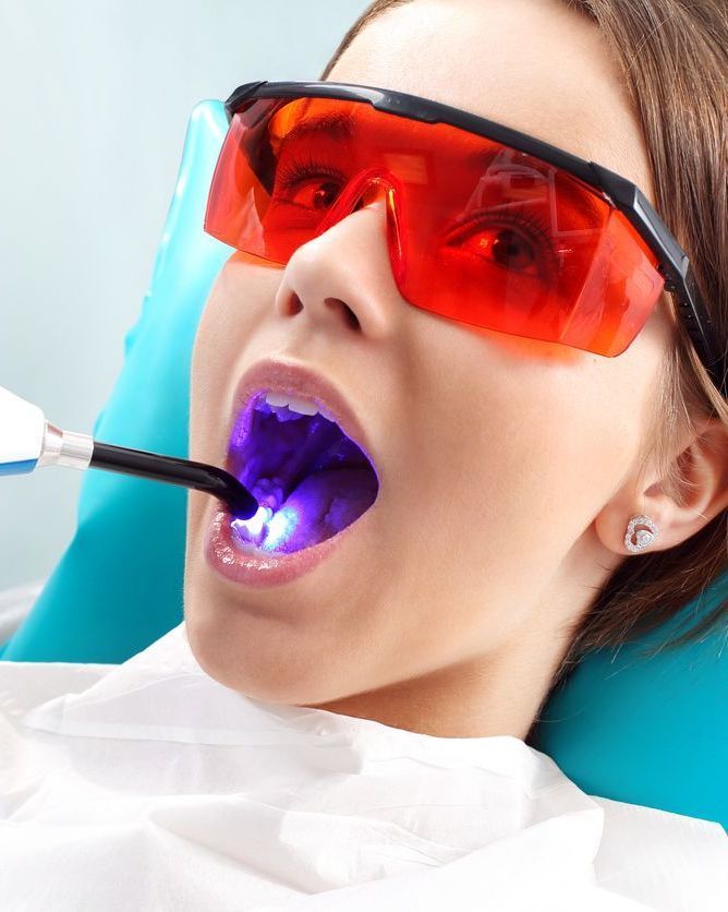 Dentist curing a patient’s tooth with blue light while she wears red protective glasses