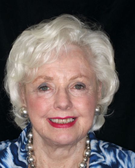 Portrait of an elderly woman with white hair, blue patterned blouse, and pearl necklace, smiling against a dark background