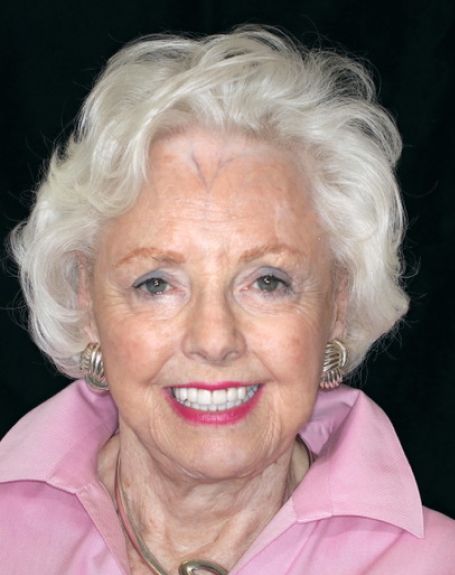 Smiling woman with short white hair wearing a pink collared shirt against a black background