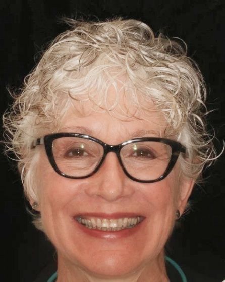 Smiling person with curly gray hair and black glasses against a dark background
