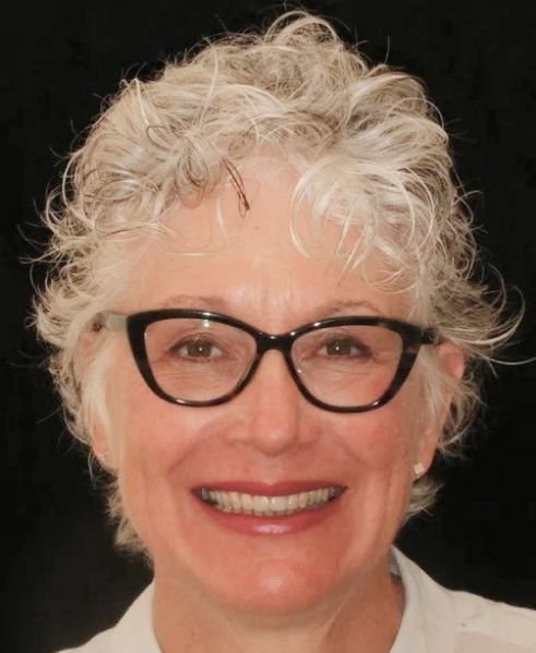 Smiling person with curly white hair and black glasses against a dark background