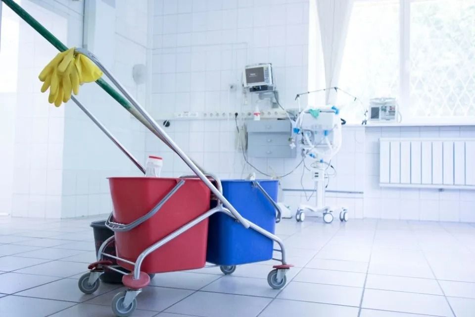 In una luminosa stanza medica bianca, un carrello per le pulizie con secchi rossi e blu e uno straccio è appoggiato su un pavimento piastrellato.