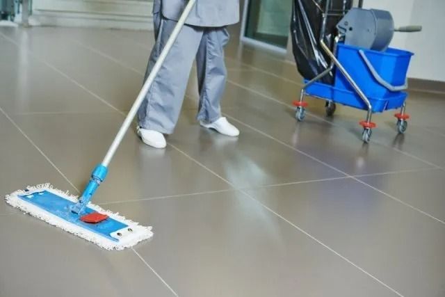 Una persona in uniforme grigia lava un ampio pavimento piastrellato accanto a un secchio blu per le pulizie con le ruote.