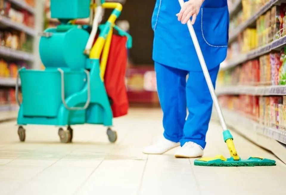 Un addetto alle pulizie in uniforme blu lava il pavimento di un supermercato accanto a un carrello per le pulizie.
