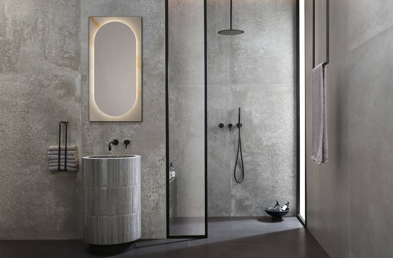 Bagno moderno con pareti grigie testurizzate, doccia, specchio ovale con luce e lavandino cilindrico.