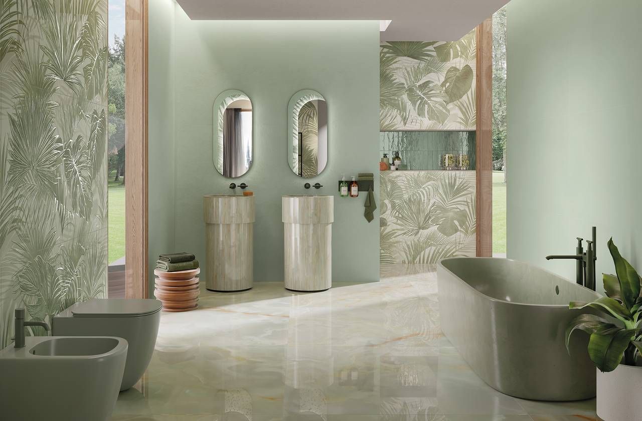 Bagno verde menta con parete decorata, due specchi ovali, lavandini su piedistallo, vasca da bagno e pianta.