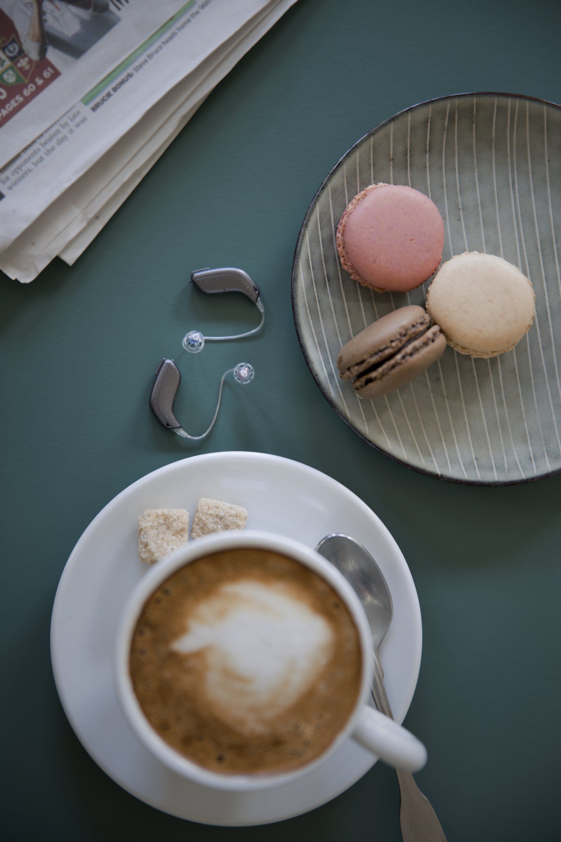 Une tasse de café sur une soucoupe à côté d'une assiette de macarons et d'une paire d'appareils auditifs.