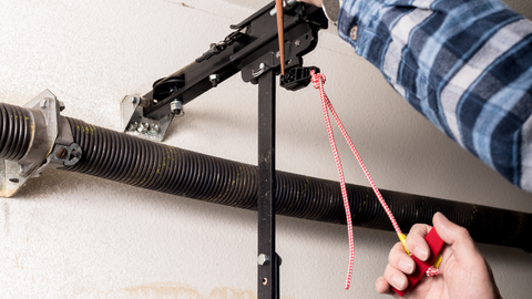 Person adjusting a garage door spring tension, using a winding bar and rope to tighten the mechanism.