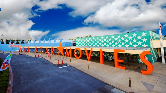 Orange “MOVIES” sign outside a teal, star-patterned building under a cloudy sky.