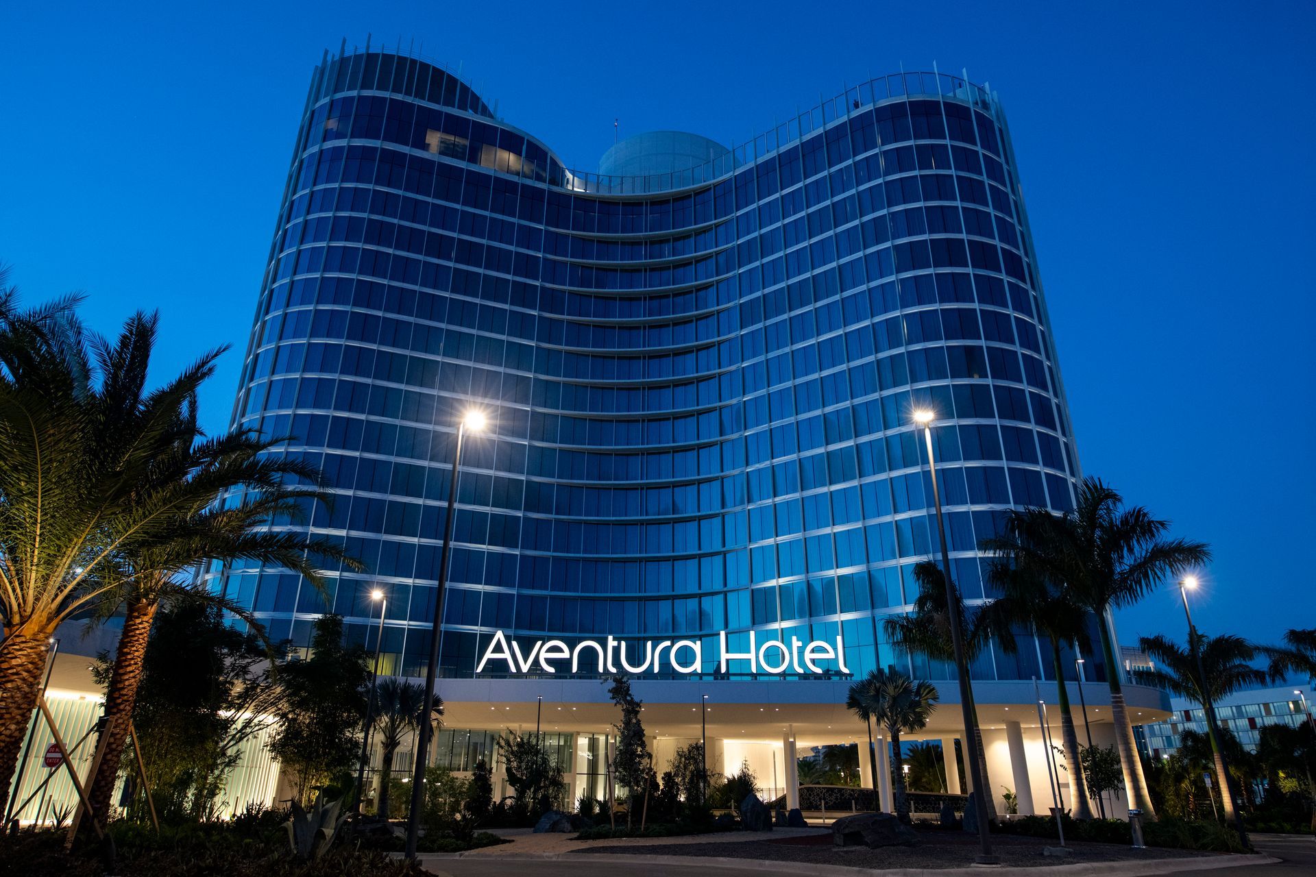 Aventura Hotel lit up at night, with a curved glass facade and palm trees in front.