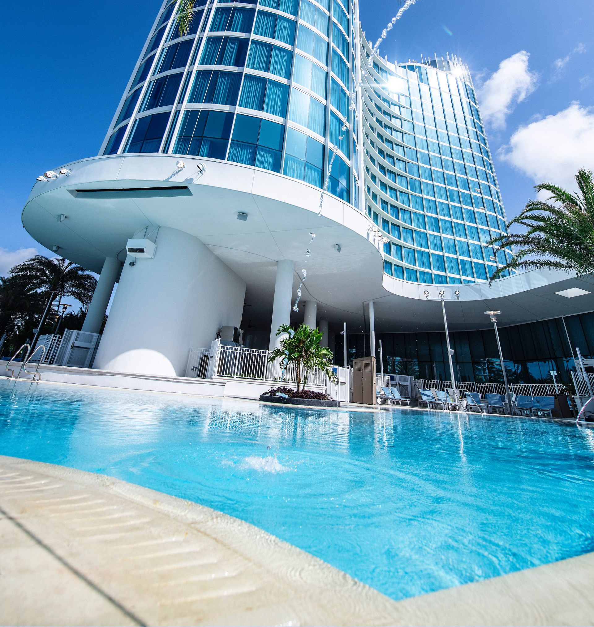 Modern glass hotel with blue pool and palm trees under a bright sky