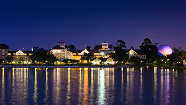 Nighttime waterfront resort with lit buildings and colorful reflections on calm water