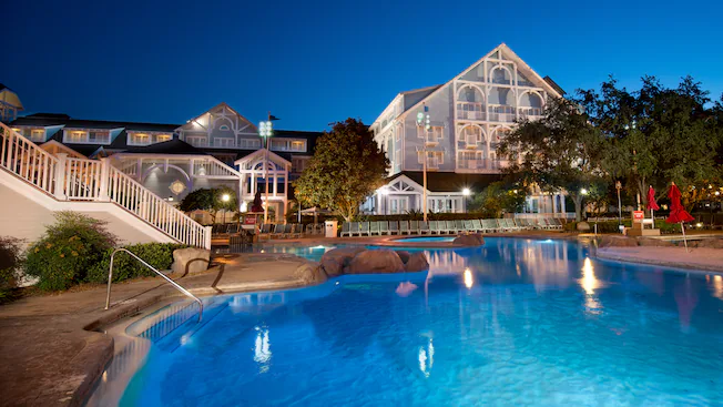 Resort pool at dusk with glowing blue water, lit buildings, and a glass atrium in the background