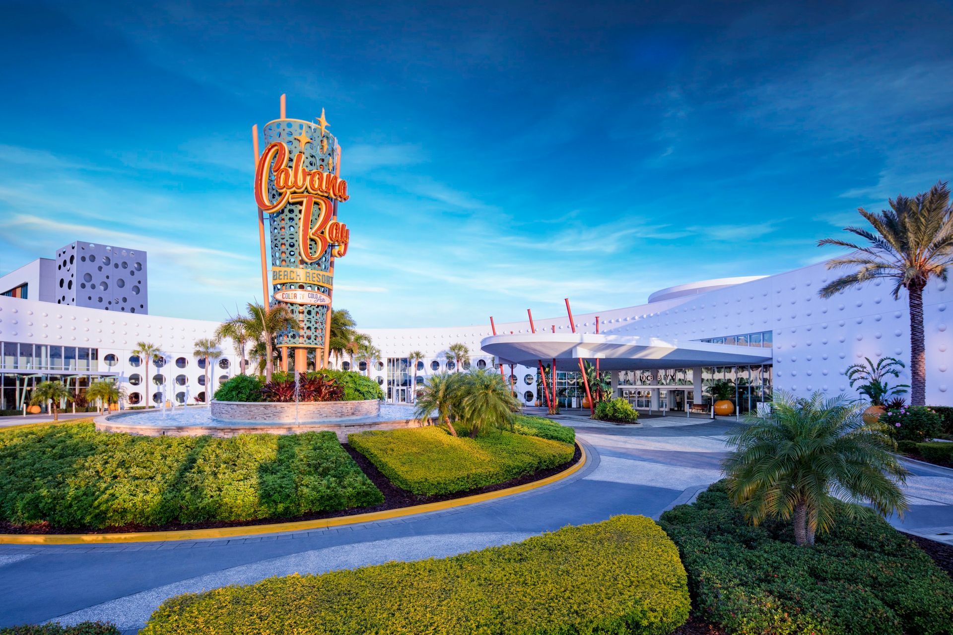 Modern white resort entrance with a colorful tower, palm trees, and a landscaped driveway at dusk
