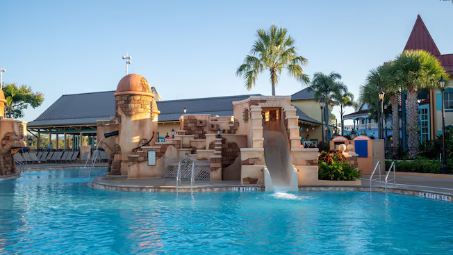 Resort pool with a stone play structure and waterslide under palm trees at sunset