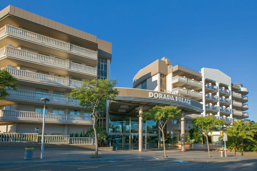 Modern hotel entrance with balconies, glass canopy, and palm trees under a clear blue sky