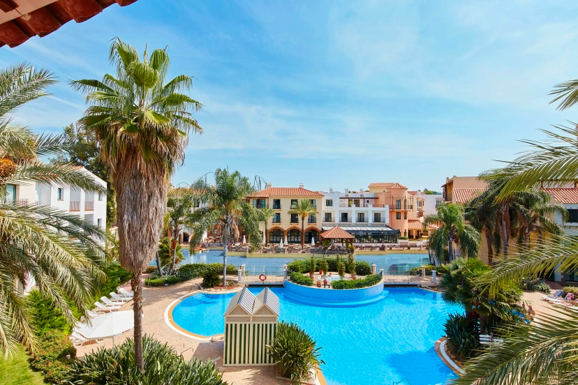 Resort pool with palm trees, blue water, and Mediterranean-style buildings under a sunny sky