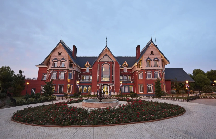 Red-brick mansion with ornate gables, a circular flower bed, and a fountain in front at dusk
