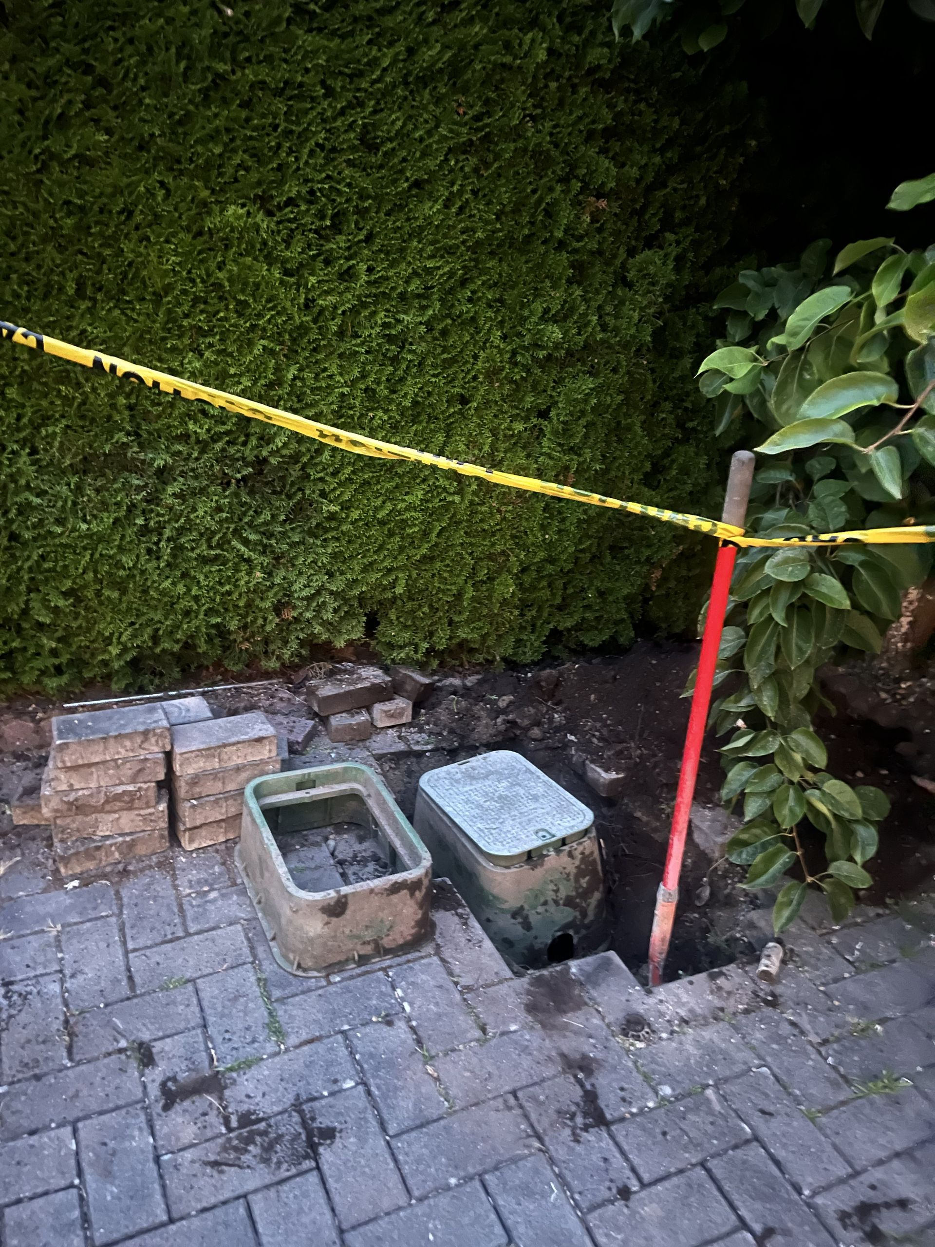 Cordoned-off garden edge with yellow tape, red post, brick paving, and two small containers near a hedge