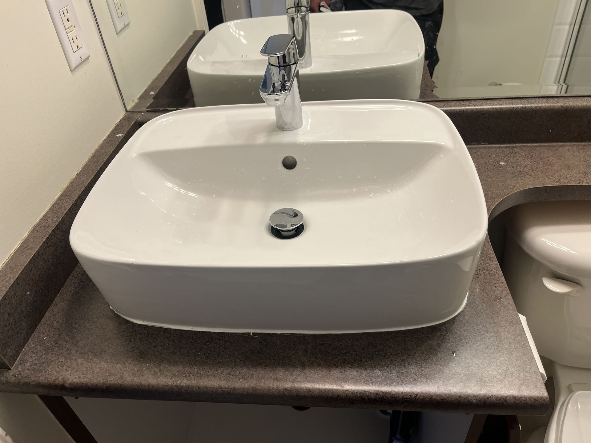 Modern white bathroom sink with chrome faucet on a dark countertop, seen in a mirror reflection.