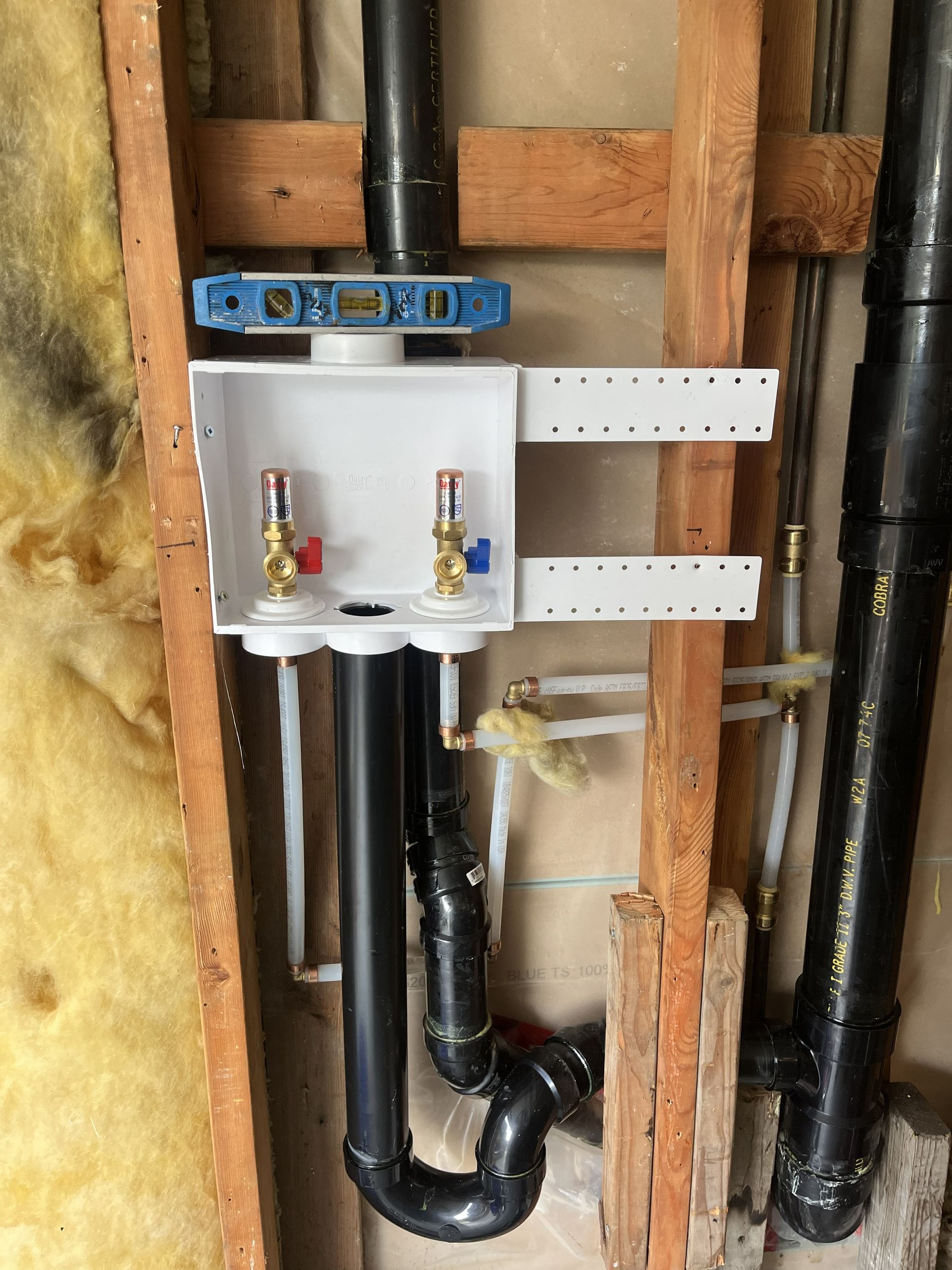 Plumbing wall with white water meter manifold, black pipes, and exposed wooden studs inside a renovation area