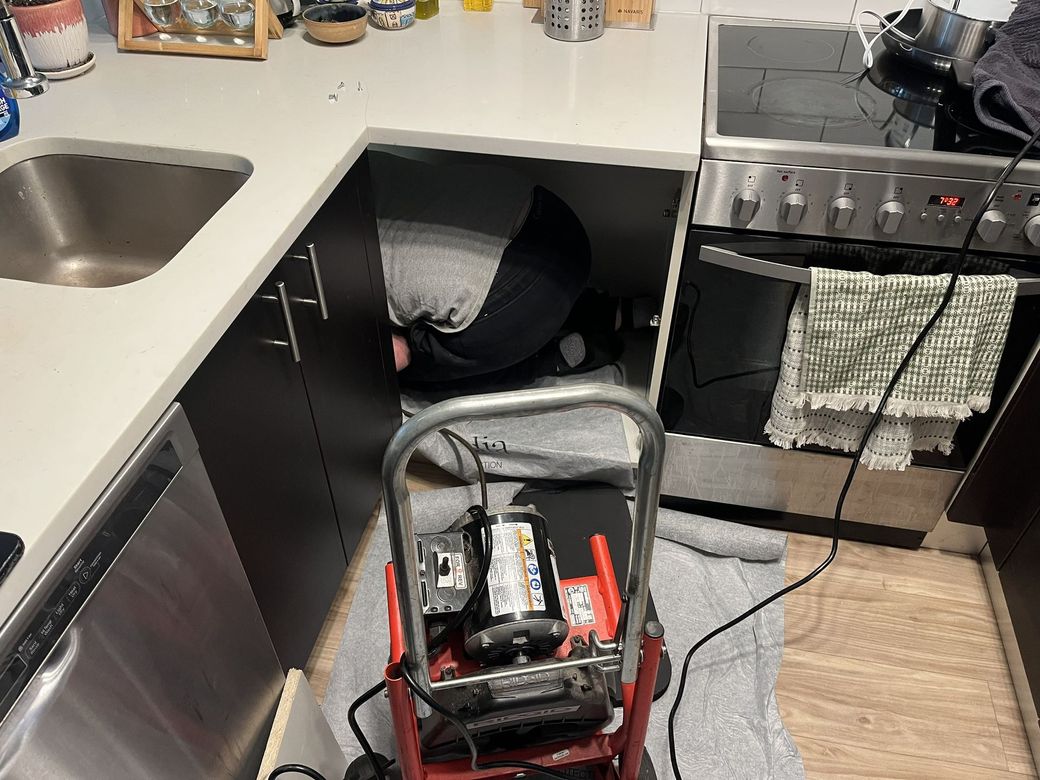Cluttered kitchen with a red wheeled machine in the foreground and a person crouched under the counter.