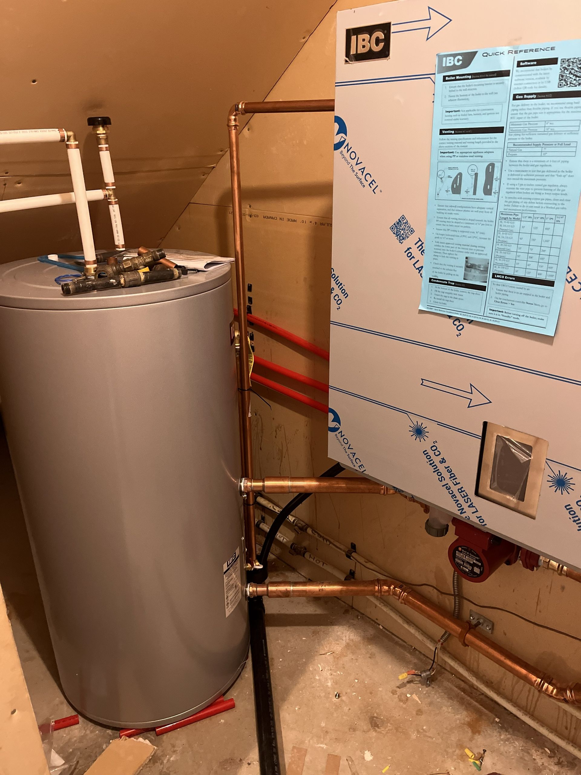Water heater and exposed plumbing in a utility room next to a beige insulated panel with posted notices.
