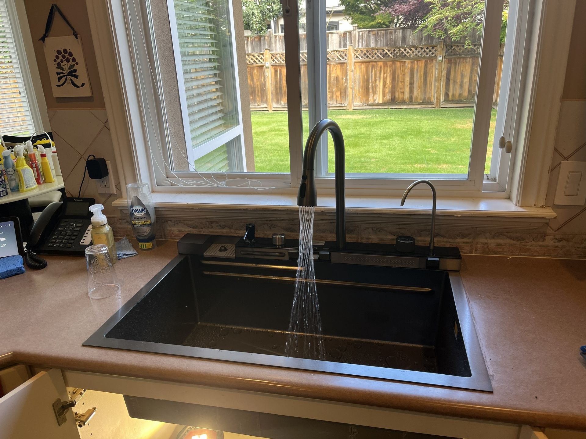 Kitchen sink with running faucet under a window, overlooking a backyard lawn and wooden fence.