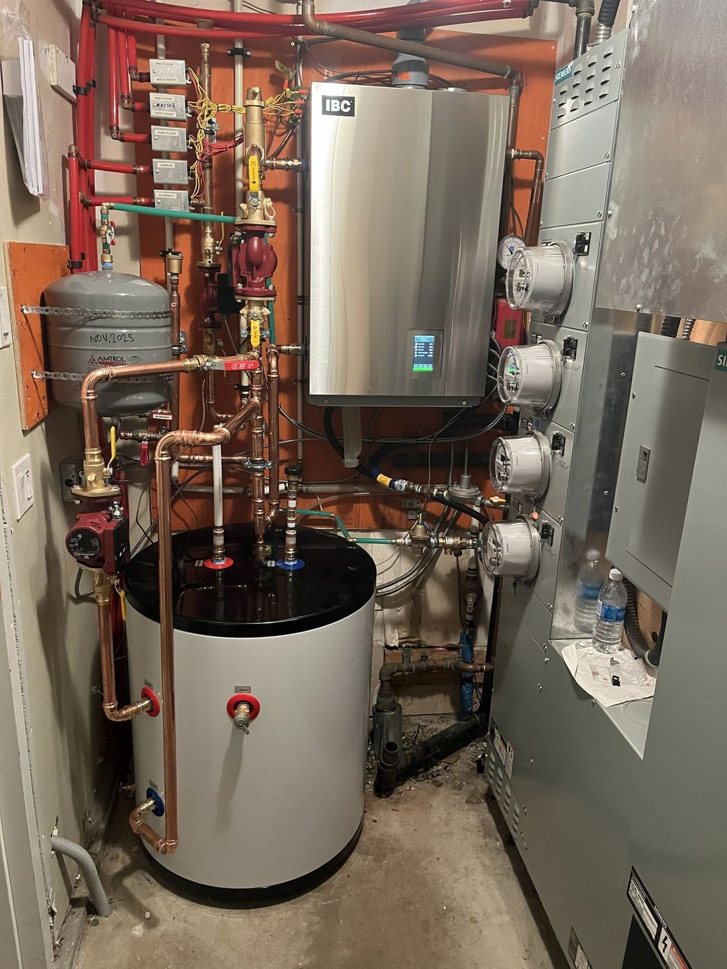 Mechanical room with boilers, pipes, and a large white tank beside a wall-mounted unit.