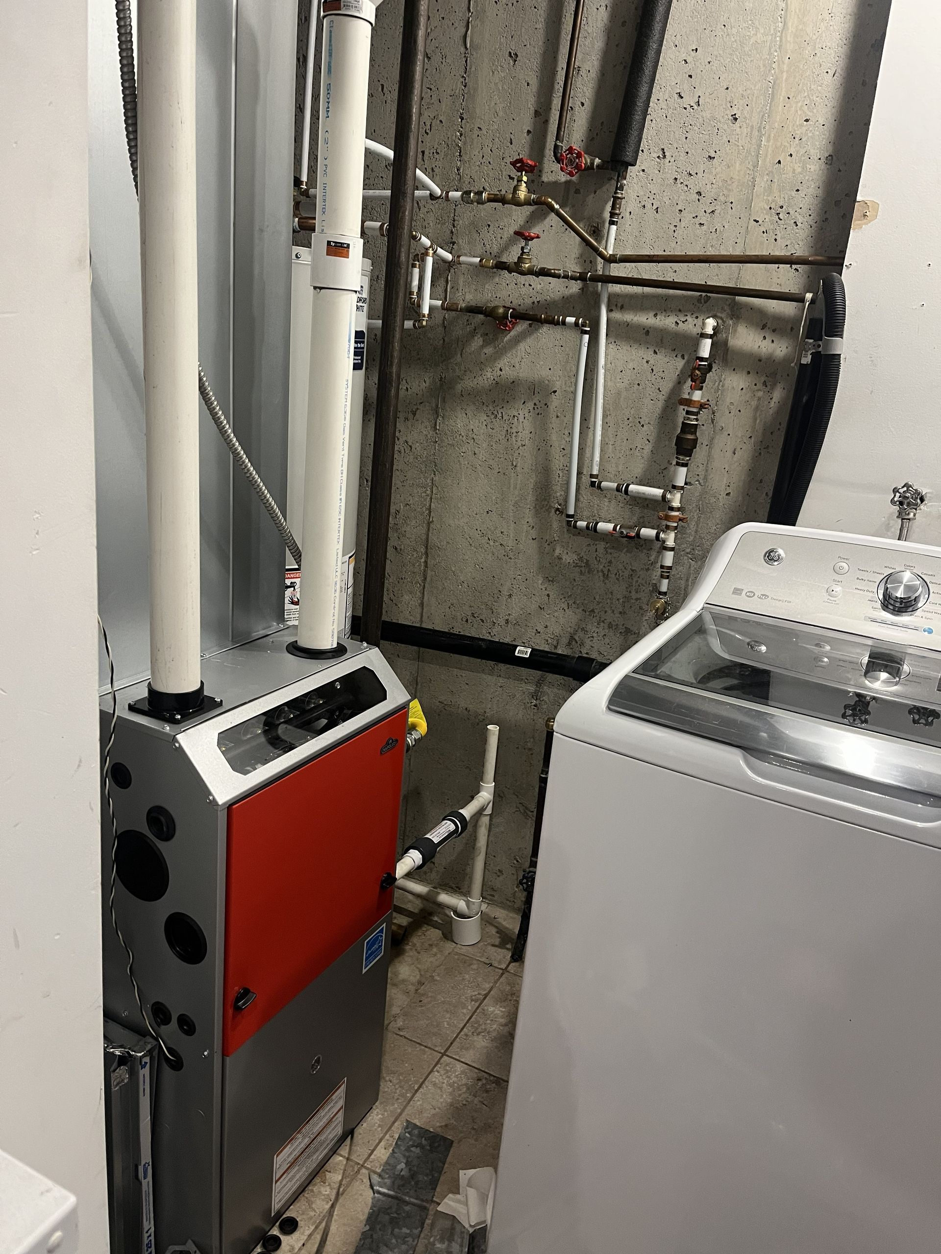 Laundry room with a washer, dryer, and red boiler units beside exposed pipes on a concrete wall