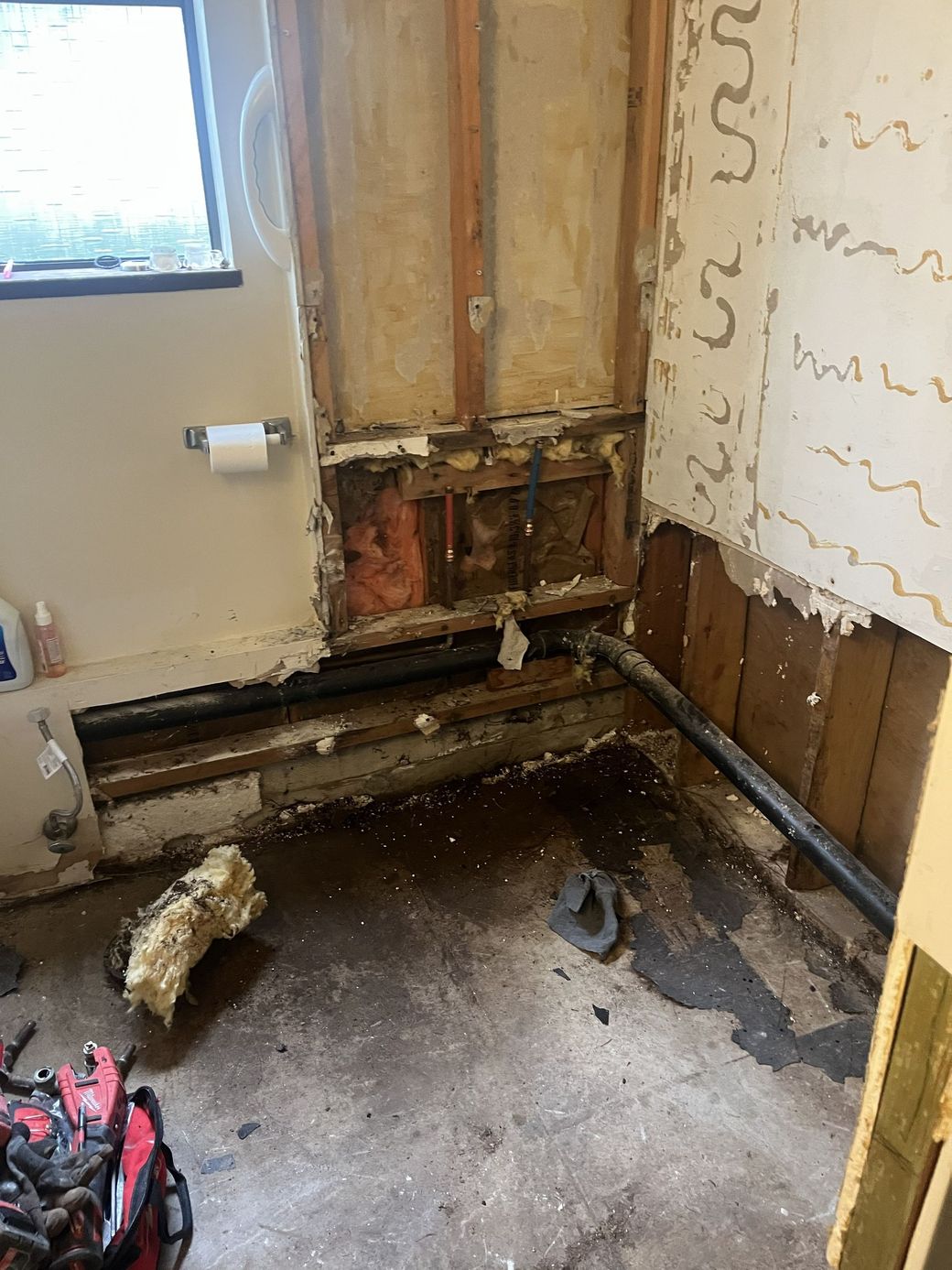 Damaged bathroom wall and floor with exposed pipes and debris during renovation