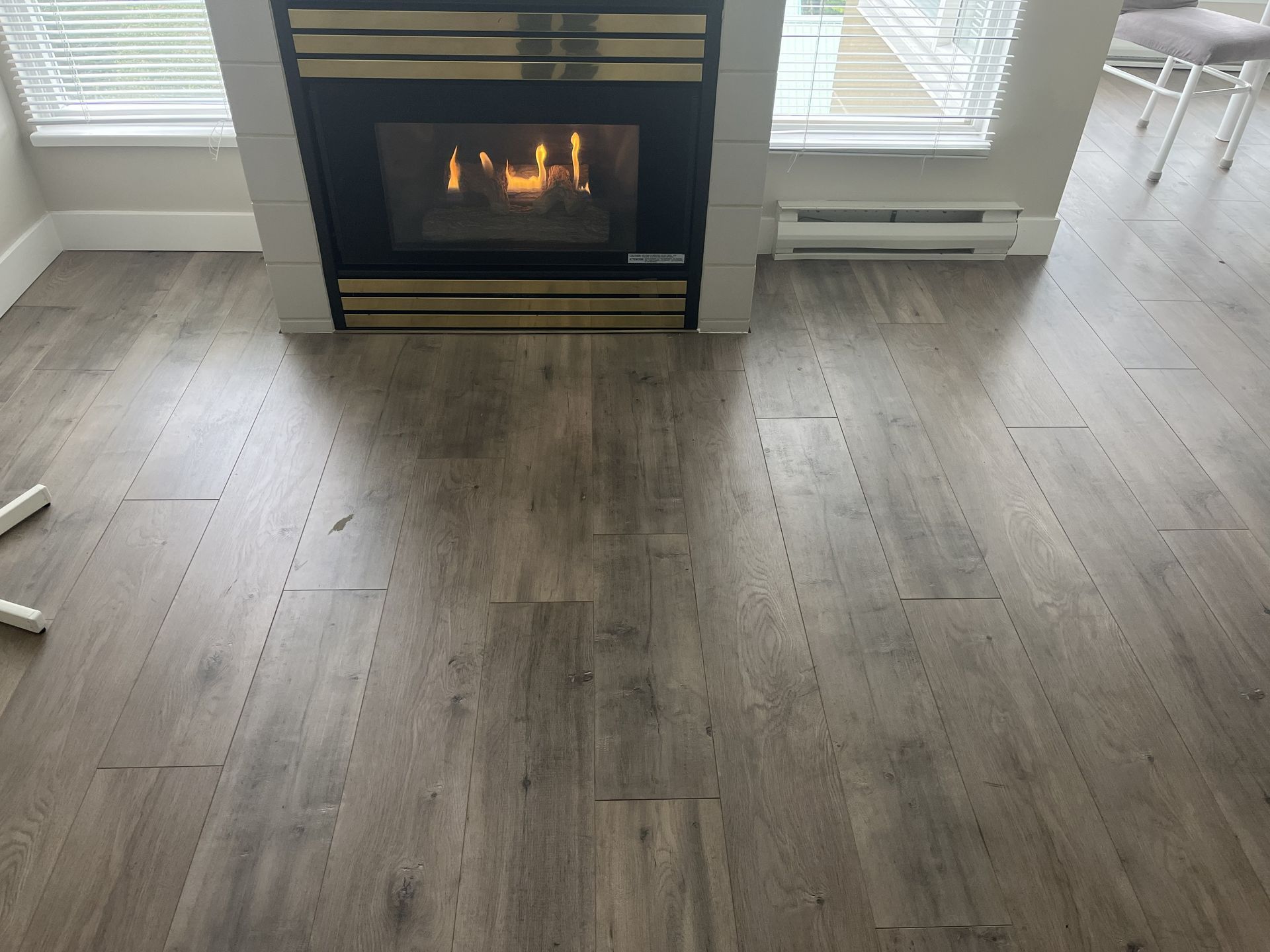 Modern living room with a lit fireplace, gray wood floor, and white walls near windows.