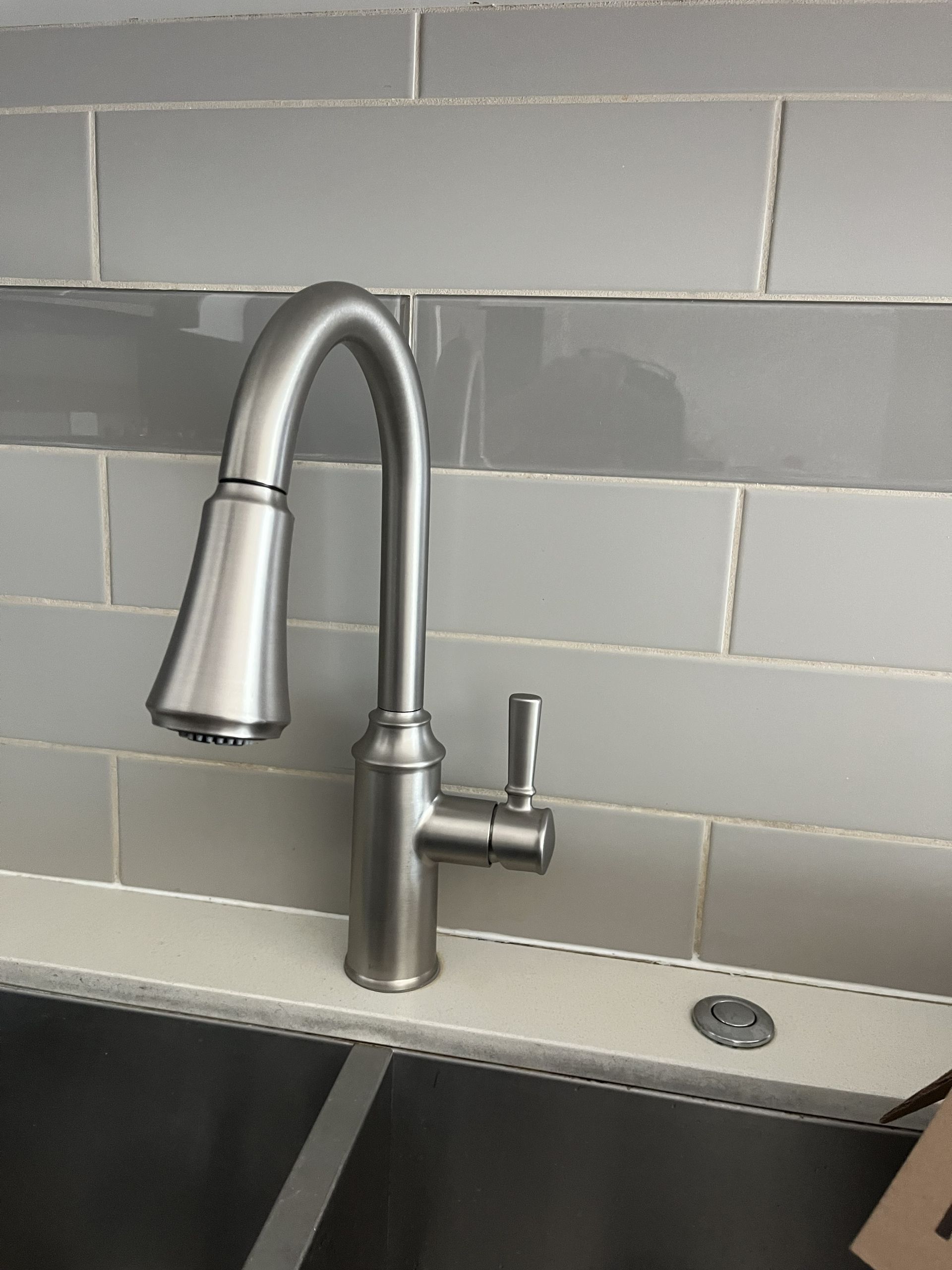 Chrome kitchen faucet beside a sink with gray tile backsplash and soap dispenser.
