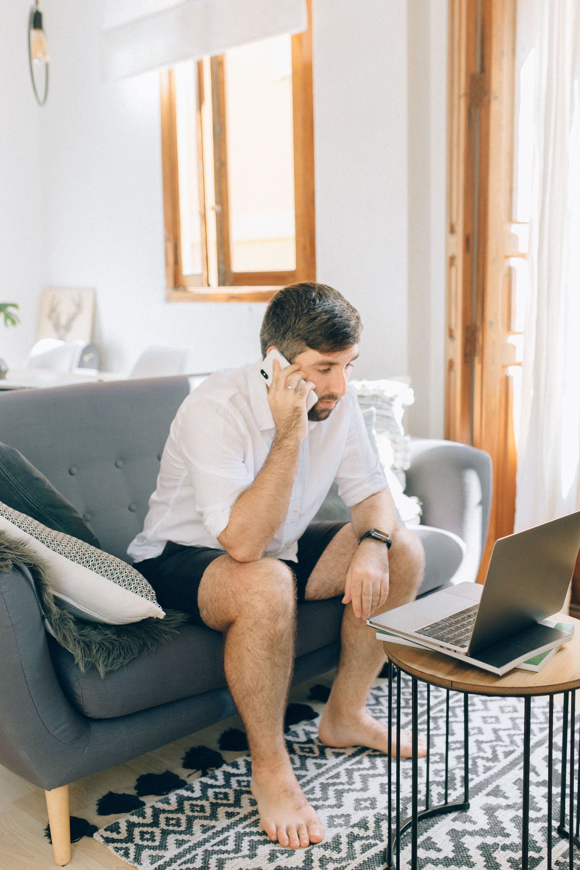 Man sitting on a gray sofa, on a phone call beside a laptop in a bright living room