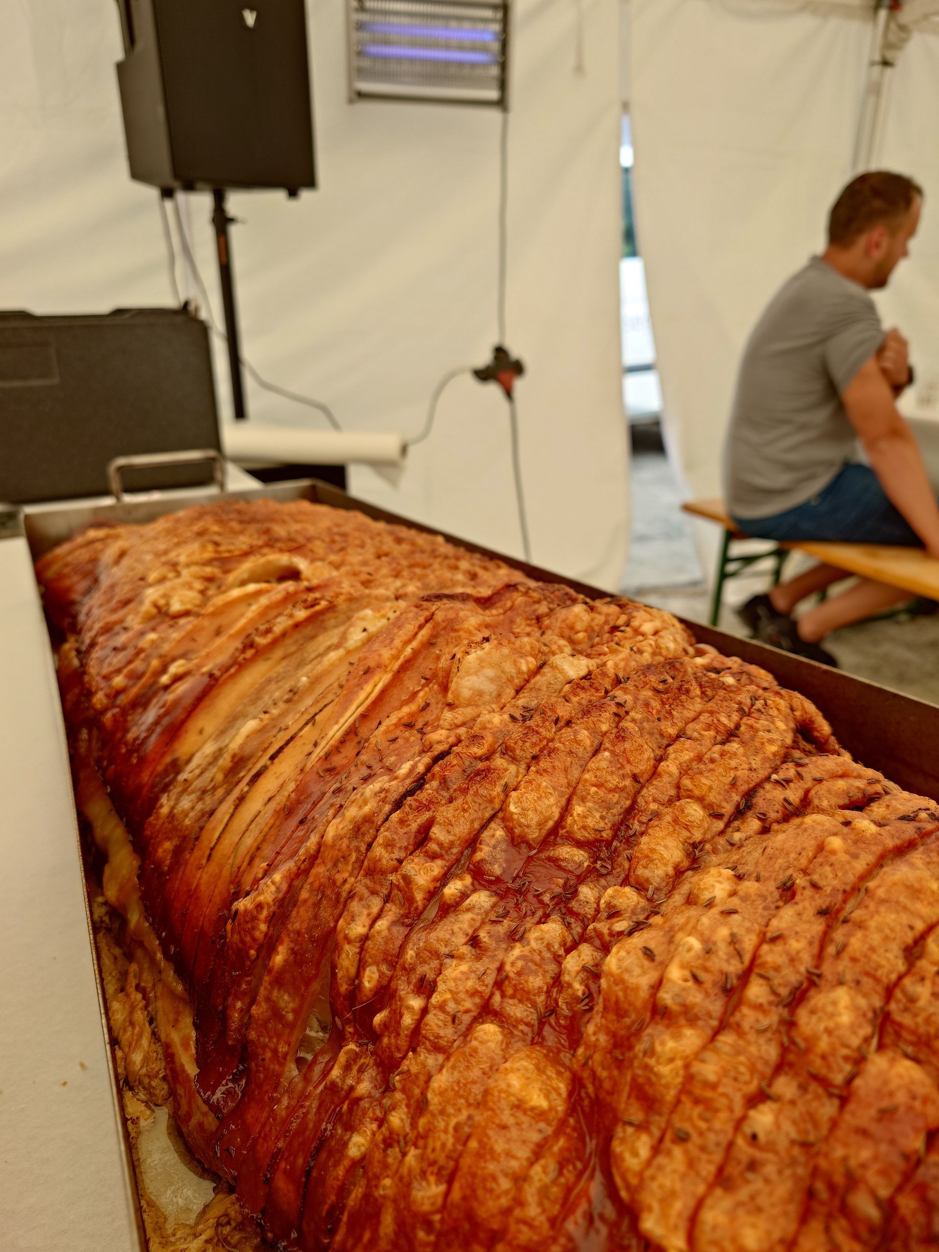 Nahaufnahme eines Spanferkels am Spieß auf einer Veranstaltung im Freien, im Hintergrund ein sitzender Mann.