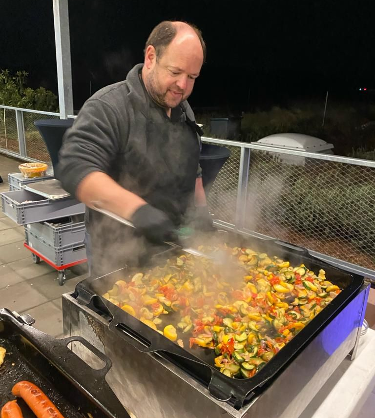 Ein Mann grillt nachts verschiedenes Gemüse auf einer großen Grillplatte im Freien, während Dampf aufsteigt.