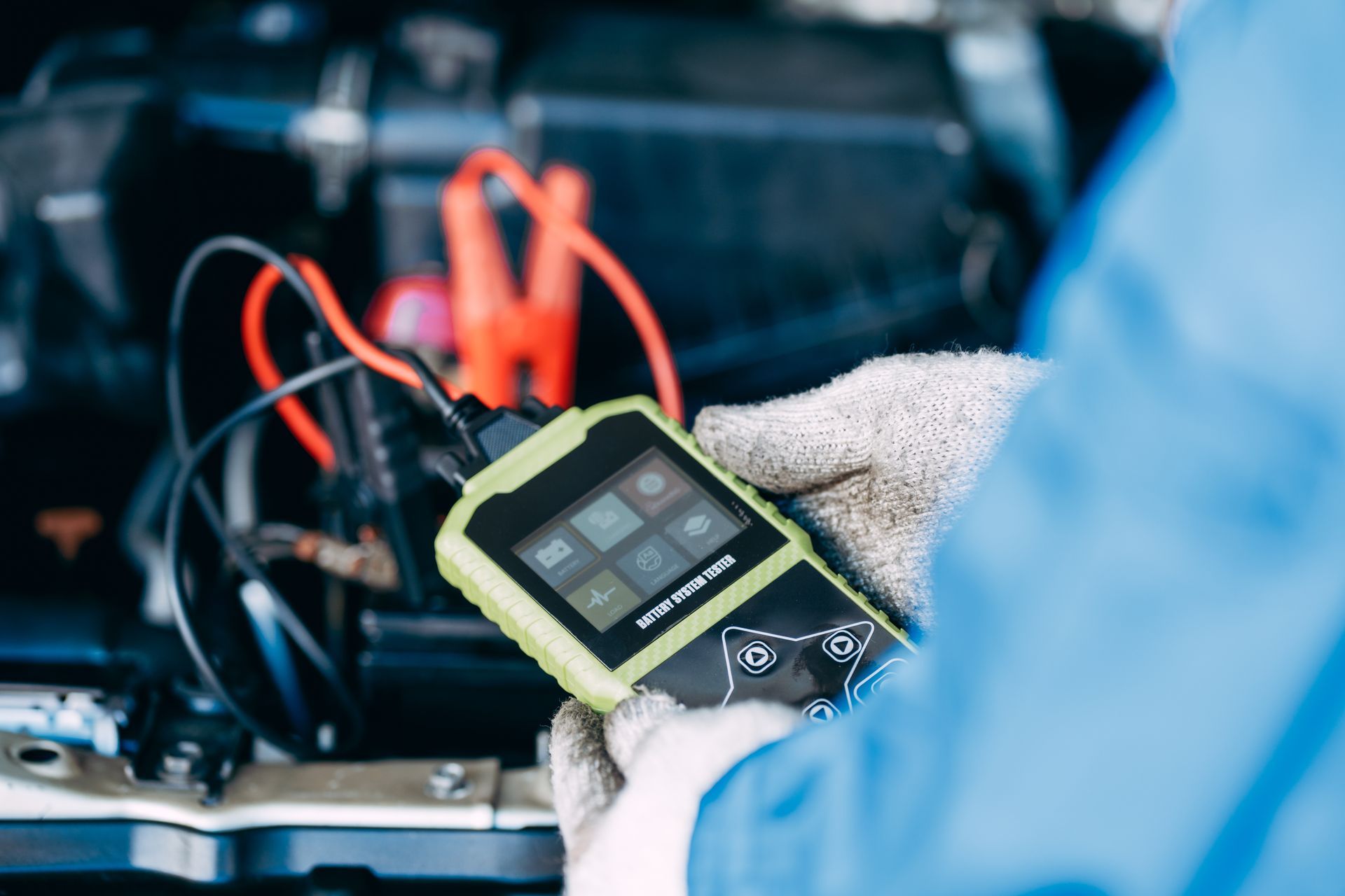 Gloved technician holding a diagnostic scanner near a car battery with red and black cables attached.