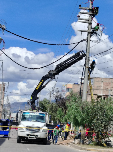 Camión de servicios públicos con grúa levantando un poste de luz en una calle, con trabajadores debajo bajo un cielo nublado.