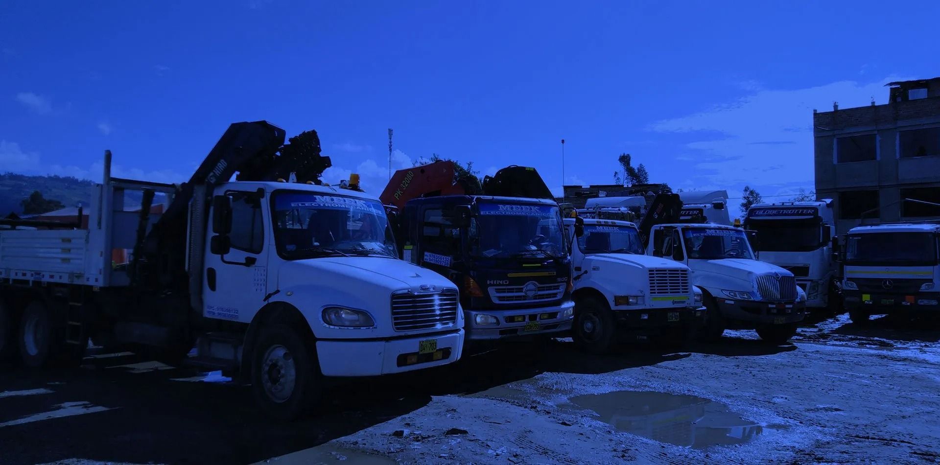 Fila de grúas y camiones de servicios públicos estacionados en un terreno nevado al anochecer.