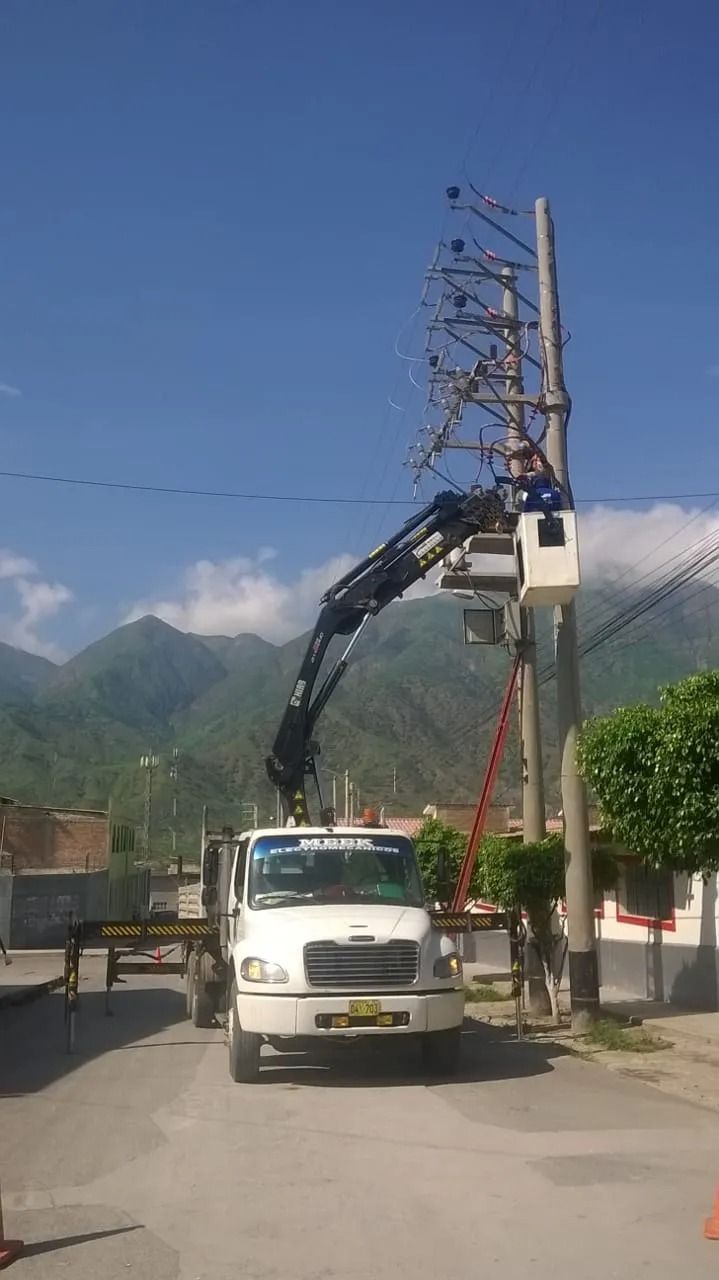 Una grúa montada sobre un camión levanta una alta torre metálica en una carretera con montañas al fondo.