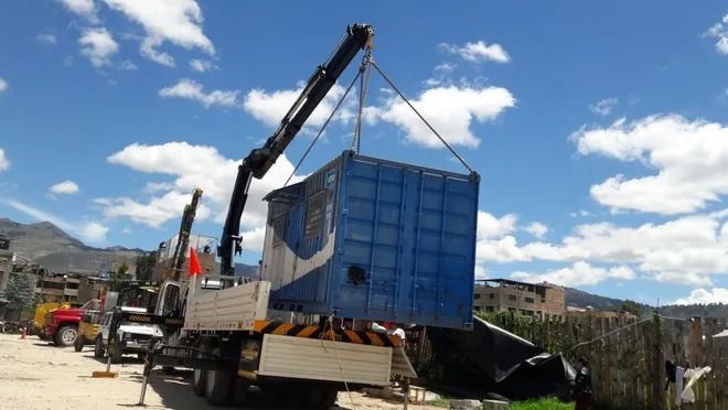 Una grúa eleva un gran contenedor azul sobre un camión en una obra vial bajo un cielo soleado.