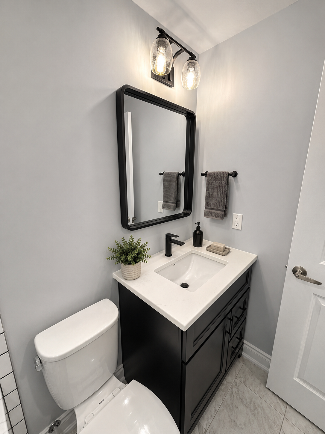 Small bathroom with white vanity, black cabinet, mirror, toilet, towel, and wall light.