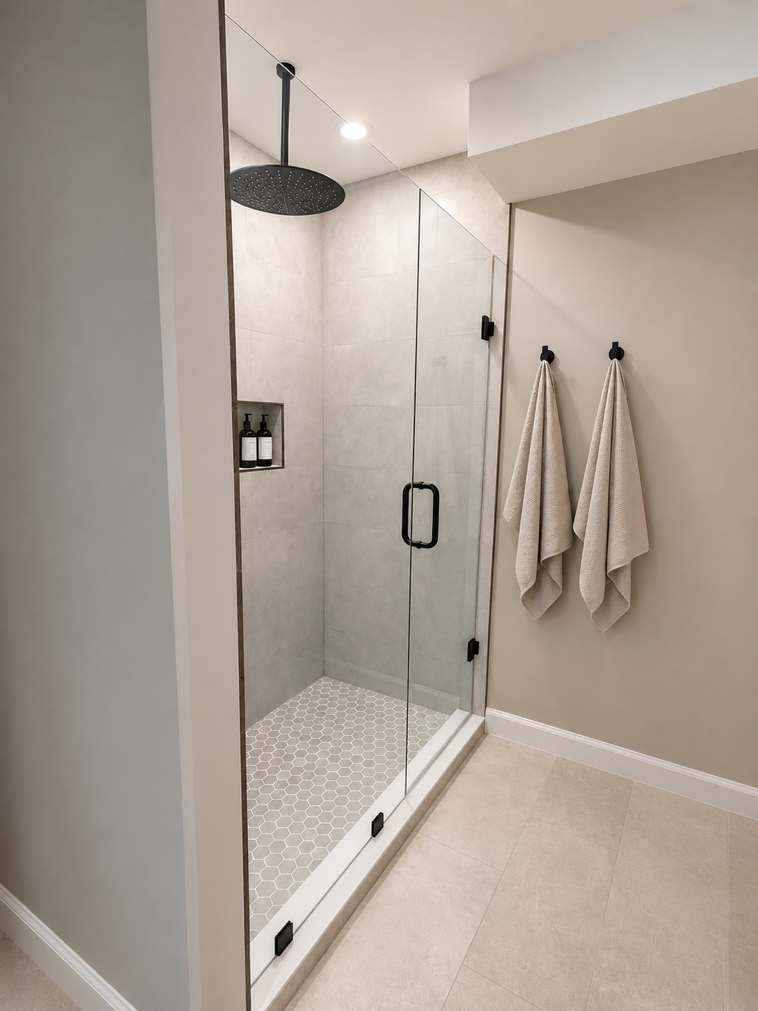 Modern shower with glass door, black fixtures, and three towels hanging on hooks in a beige bathroom.