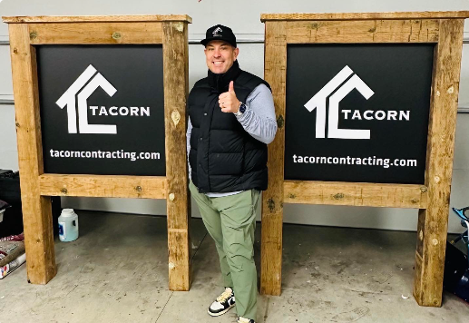 Person posing between two large Tacorn contracting signs indoors, giving a thumbs-up.