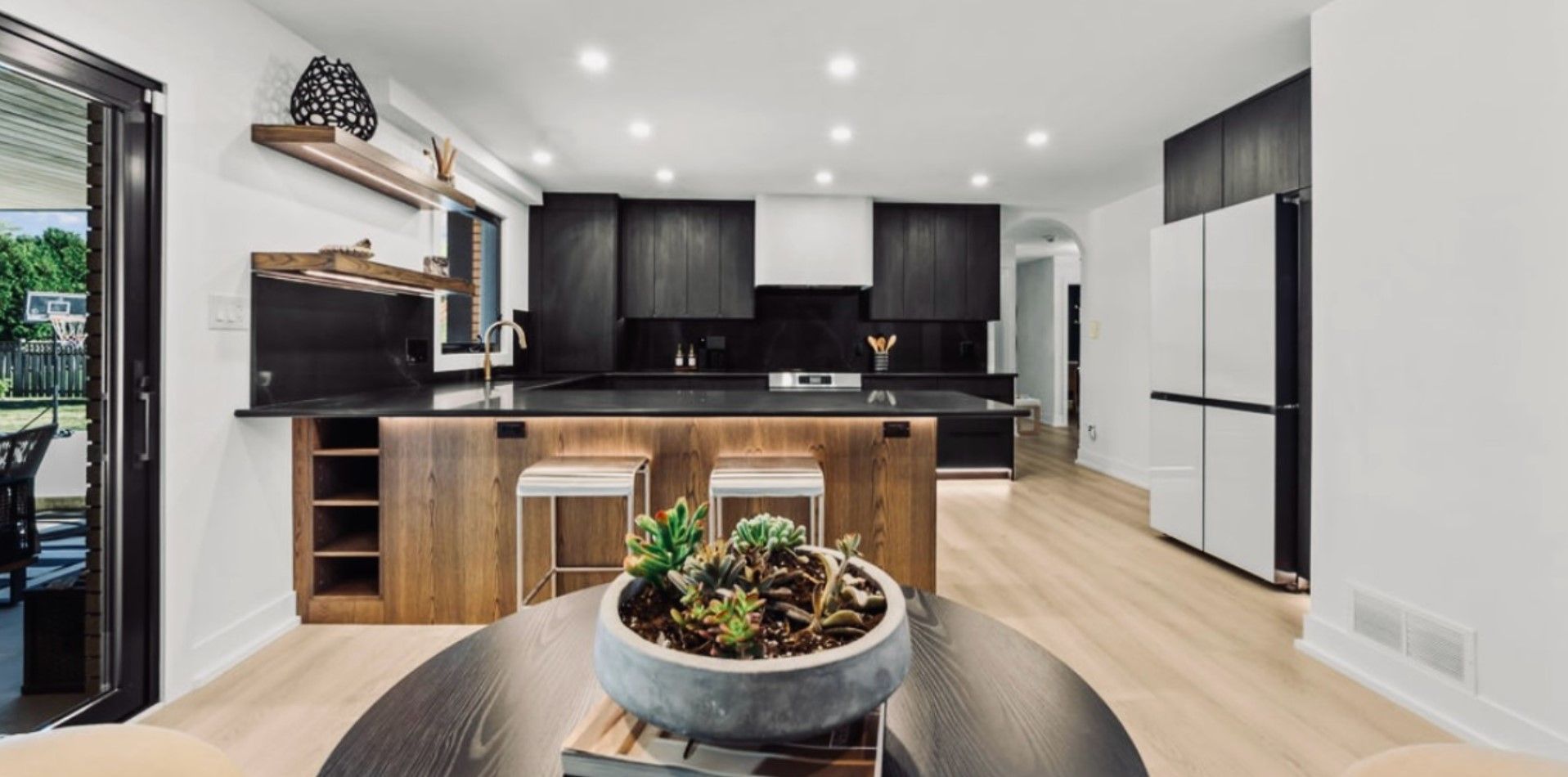 Modern open kitchen with dark cabinets, light wood floors, white island, and a bowl of succulents on a table