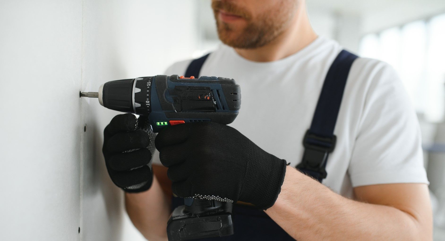 Person drilling into a wall with a cordless power drill, wearing black gloves and white shirt
