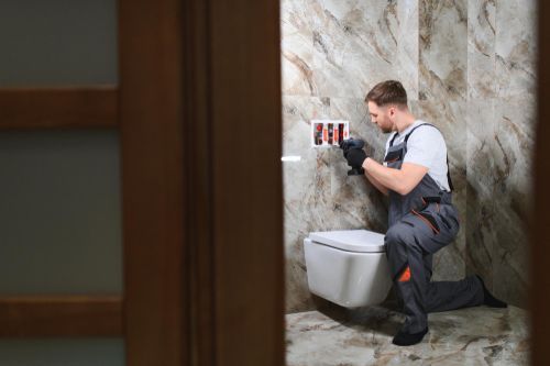 Worker kneeling beside a toilet, using a tool on the wall in a tiled bathroom.