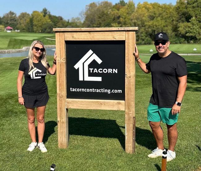 Two people standing beside a TACOM sign on a grassy golf course under sunny skies