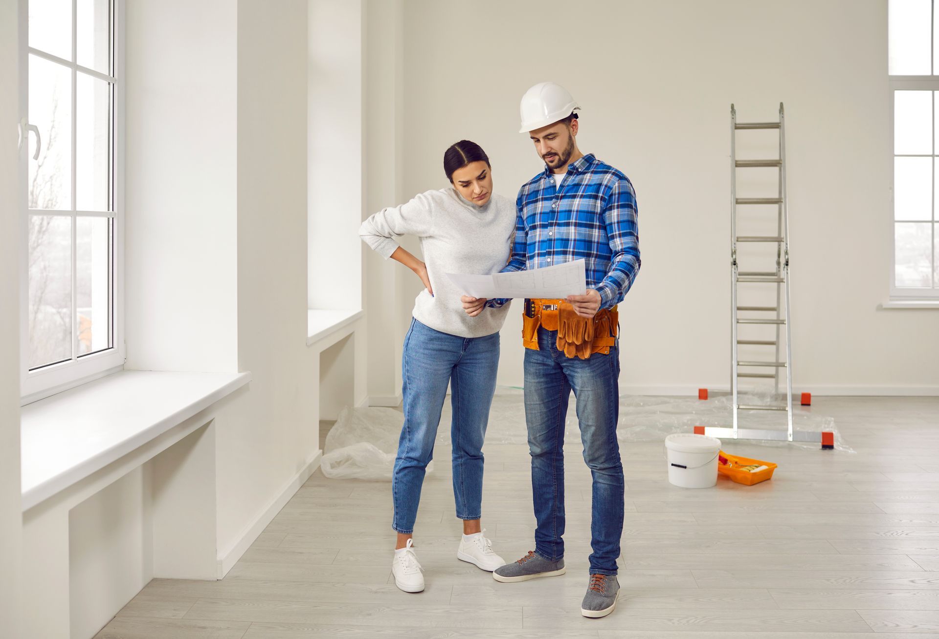 Two people reviewing a renovation plan in a bright, empty room with paint supplies and a ladder.
